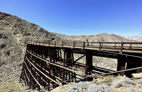 A wooden bridge spans a rocky canyon, surrounded by mountains under a clear blue sky.