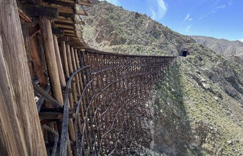 A curved wooden trestle bridge spans a rocky landscape, leading to a tunnel in the mountains under a blue sky.
