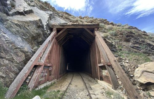 A wooden tunnel entrance set in rocky terrain, leading into darkness with a railway track visible inside.