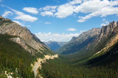 A scenic view of a winding road through a lush green valley surrounded by towering mountains and a blue sky with clouds.