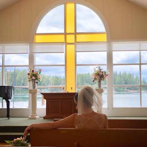 A bride sits in a serene chapel, with a large cross and floral arrangements in the background, overlooking a scenic view.