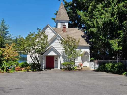 A white church with a steeple surrounded by trees and a clear blue sky, set in a parking lot.