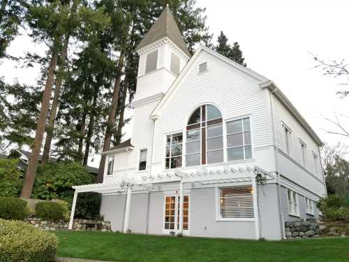 A large white house with a tower, surrounded by trees and a green lawn, featuring large windows and a patio area.