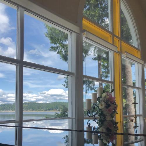 A serene view through large windows, showcasing a lake and trees, with a floral arrangement and candles in the foreground.
