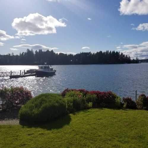 A serene waterfront scene with a boat docked near lush greenery and colorful flowers under a partly cloudy sky.