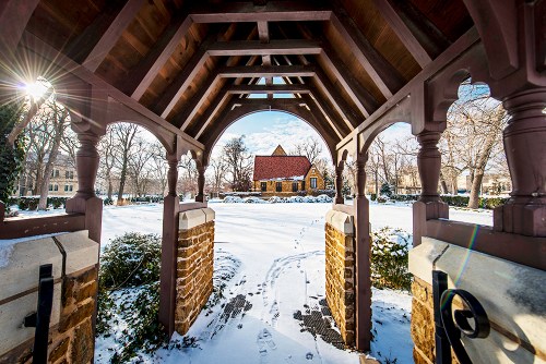 A snow-covered landscape viewed through a wooden archway, leading to a charming house in the distance.