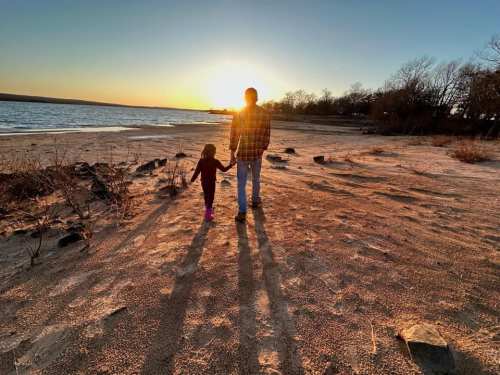 A person and a child walk hand in hand along a sandy beach at sunset, casting long shadows.