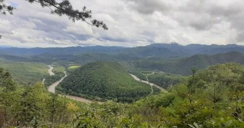A panoramic view of rolling green hills and a winding river under a cloudy sky.