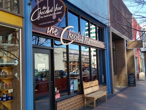 Exterior of "The Chocolate Bar," a shop featuring handcrafted chocolates, with a wooden bench outside.