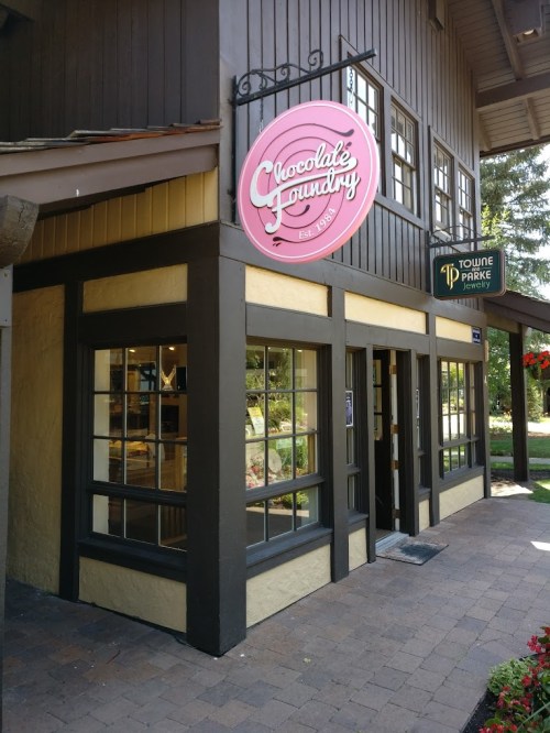 Exterior of a shop called "Chocolate Foundry," featuring a pink sign and large windows, surrounded by greenery.