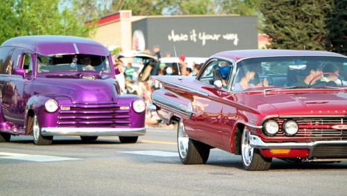 Two classic cars, a purple truck and a red convertible, cruising down a street during a car show.