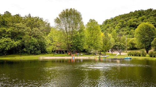 A serene lake surrounded by lush greenery, with a cabin and kayaks visible on the shore under a cloudy sky.