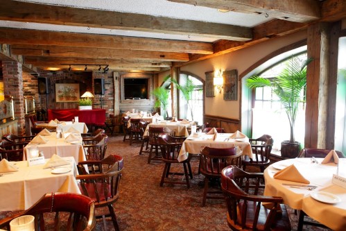 Cozy restaurant interior with wooden beams, tables set with white linens, and plants near large windows.