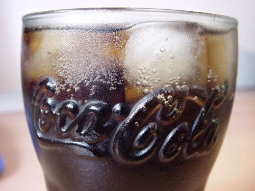Close-up of a glass of cola with ice, featuring the Coca-Cola logo and bubbles rising in the drink.