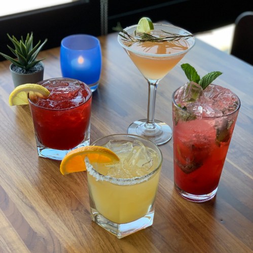 Four colorful cocktails on a wooden table, featuring garnishes like lemon, lime, and mint, with a candle in the background.