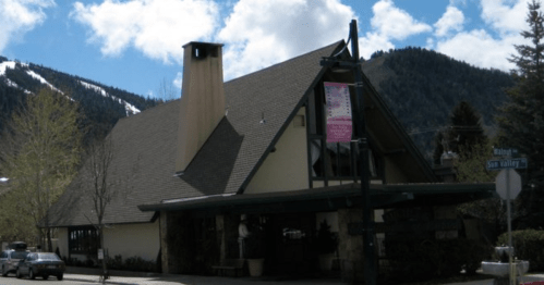A mountain lodge-style building with a sloped roof, surrounded by trees and mountains under a partly cloudy sky.