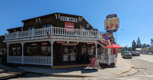 A charming candy store with a wooden exterior, featuring a sign and outdoor seating, located on a busy street.
