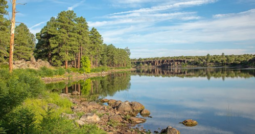 A serene lake surrounded by lush green trees and rocky shores under a clear blue sky.