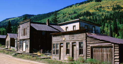 Abandoned wooden buildings line a dirt road, surrounded by lush green mountains under a clear blue sky.