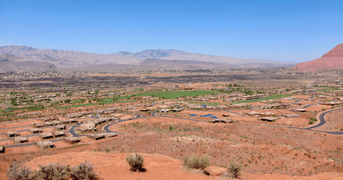 A panoramic view of a desert landscape with scattered homes, green fields, and distant mountains under a clear blue sky.