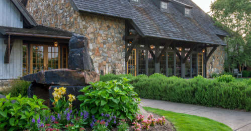 A stone-clad building with large windows, surrounded by colorful flowers and greenery, set in a serene landscape.