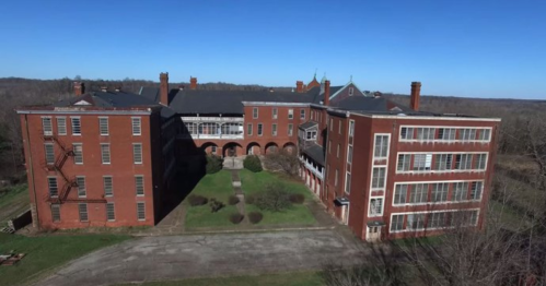 Aerial view of a large, abandoned red brick building complex surrounded by trees and a clear blue sky.