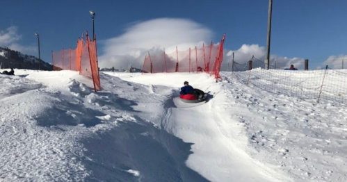 A person sleds down a snowy hill, surrounded by orange safety fencing and a clear blue sky.