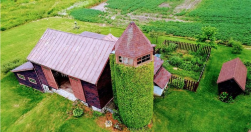 Aerial view of a rustic barn with a green, ivy-covered silo surrounded by lush fields and a small garden.