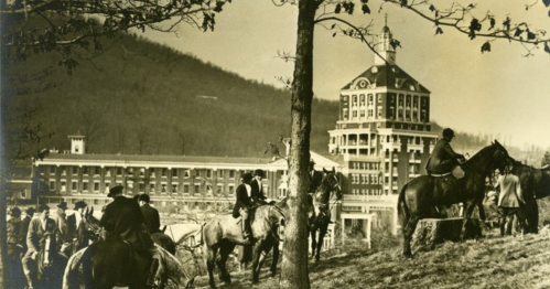 Historic image of people on horseback near a large building, with mountains in the background and trees in the foreground.
