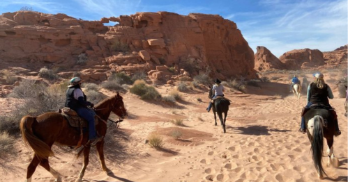 A group of people riding horses through a sandy desert landscape with red rock formations and blue skies.