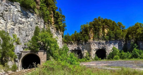 Two dark tunnels set in rocky cliffs, surrounded by lush greenery and a clear blue sky.