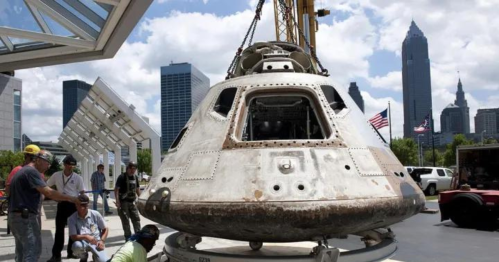 A spacecraft being lifted by a crane, with city skyscrapers in the background and people observing nearby.