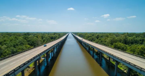 Aerial view of a long highway bridge spanning over a wide river, surrounded by lush greenery under a clear blue sky.