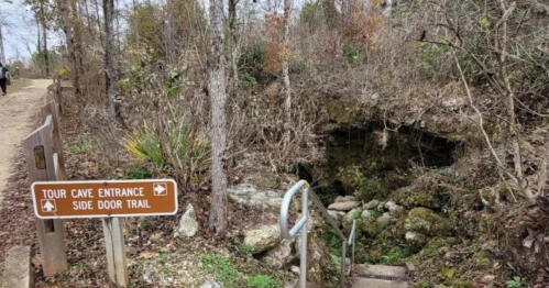 A sign points to a cave entrance and side door trail, surrounded by trees and rocky terrain. Steps lead down into the cave.