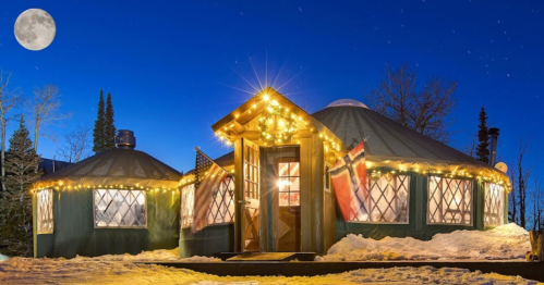 A cozy yurt adorned with lights, surrounded by snow under a bright moonlit sky. Pine trees in the background.
