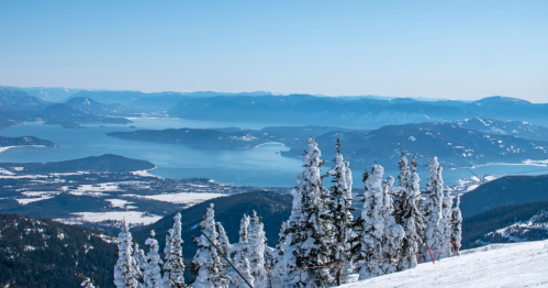 Snow-covered trees overlook a serene lake surrounded by mountains under a clear blue sky.