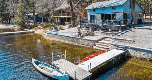 A serene lakeside view featuring a house, dock, and kayaks on calm water surrounded by trees.