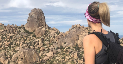 A hiker with a ponytail and headband gazes at rocky terrain and a large boulder in a scenic outdoor landscape.