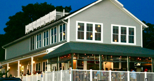 A two-story restaurant with large windows, outdoor seating, and a rooftop deck, illuminated at dusk.