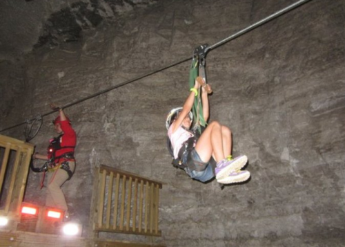 A person zip-lining inside a cave, with another person in safety gear on a platform nearby.