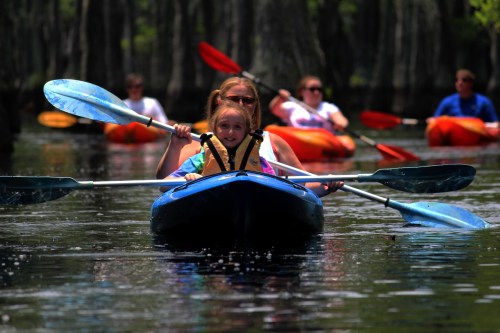A child and an adult kayak together on calm water, surrounded by others in colorful kayaks and lush greenery.