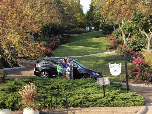 A family stands by a car in a landscaped park, surrounded by greenery and flowers, with an exit sign nearby.