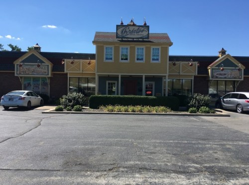 A yellow and brick restaurant building with a sign, surrounded by greenery and parked cars under a clear blue sky.