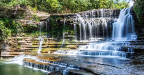 A serene waterfall cascading over rocky tiers, surrounded by lush greenery and a tranquil pool below.