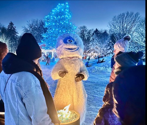 A snowy scene with a blue-lit tree, featuring a person in a yeti costume interacting with a group of people.