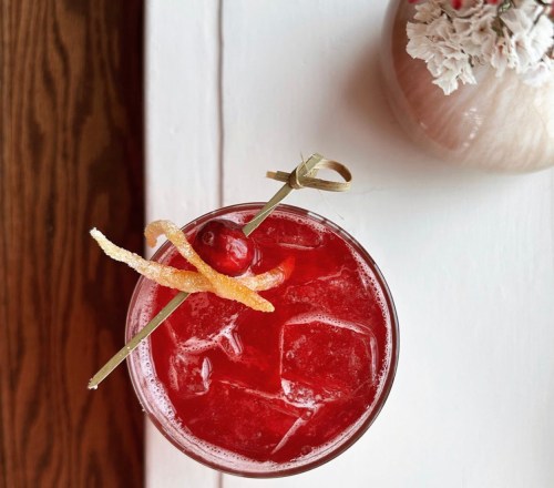 A vibrant red cocktail with ice, garnished with a cherry and citrus peel, on a white table next to a floral vase.