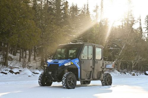 A blue all-terrain vehicle parked on snow-covered ground, surrounded by trees and sunlight filtering through.