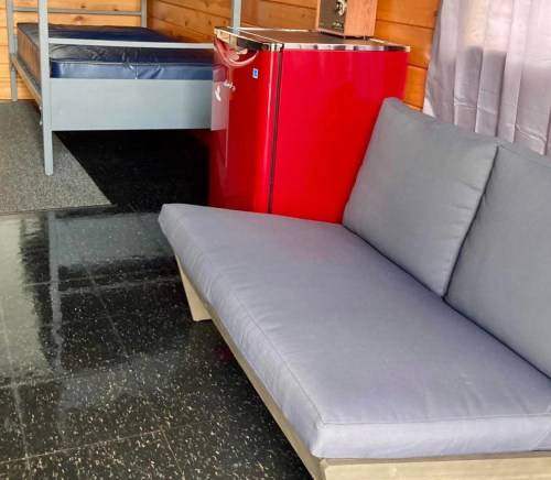A small room featuring a blue bed, a red mini fridge, and a gray couch on a black floor.