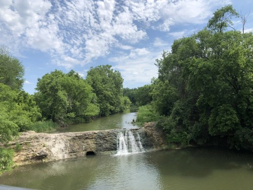 A serene river scene with a small waterfall surrounded by lush green trees and a blue sky with scattered clouds.