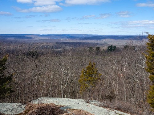 A panoramic view of a valley with bare trees and distant hills under a blue sky with scattered clouds.